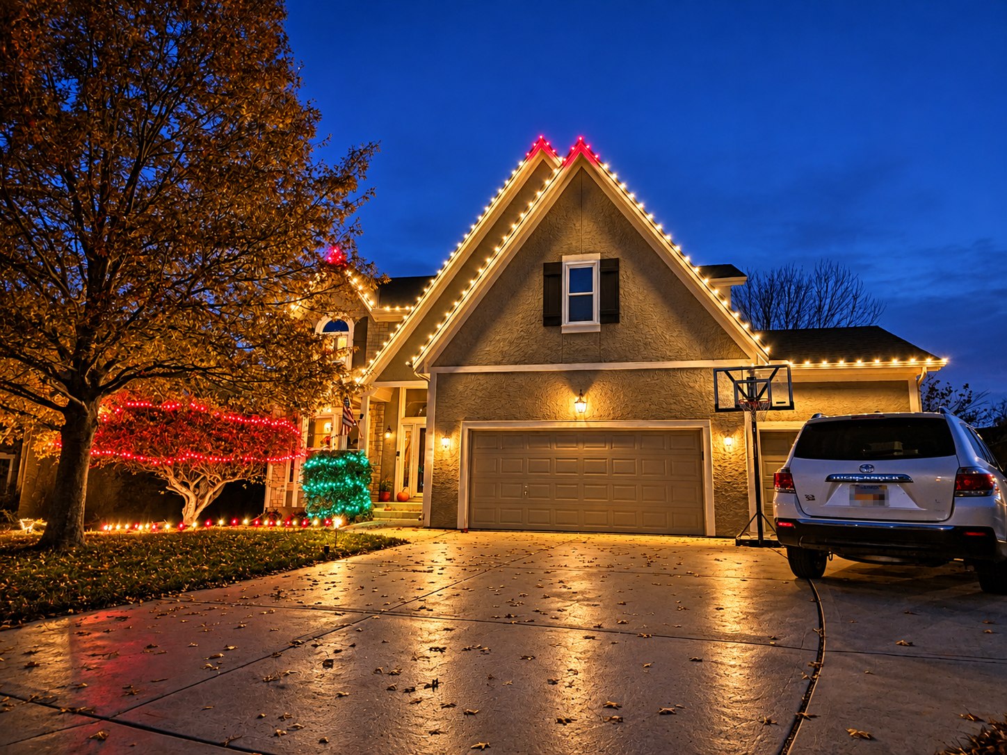 Two-story home at dusk with warm white bulbs tracing every gable and roofline, red bulbs capping the peaks, a red-lit maple and a green-lit shrub by the front steps